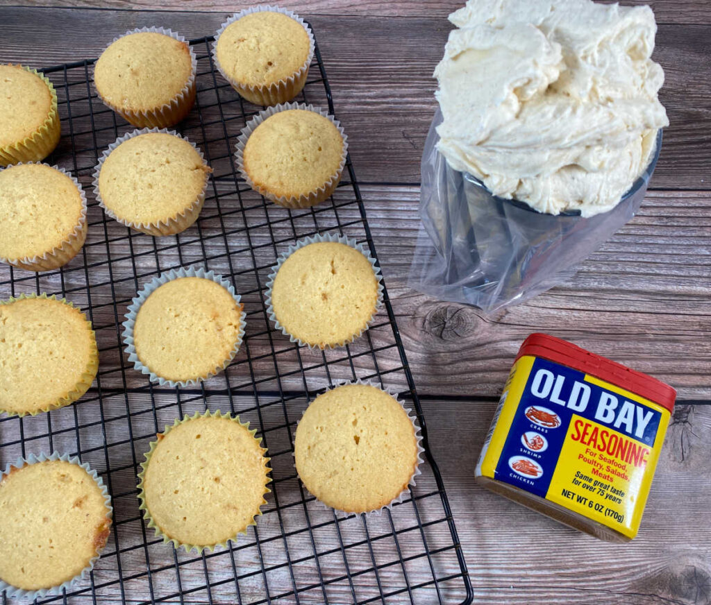 Process shot - baked cupcakes sit a on wire rack with the frosting and a can of Old Bay next to them. 