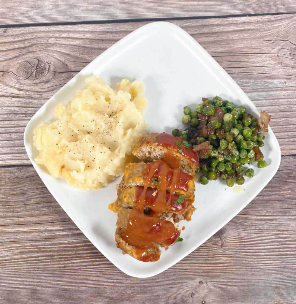 Overhead image of sliced meatloaf on a plate with mashed potatoes and peas. 