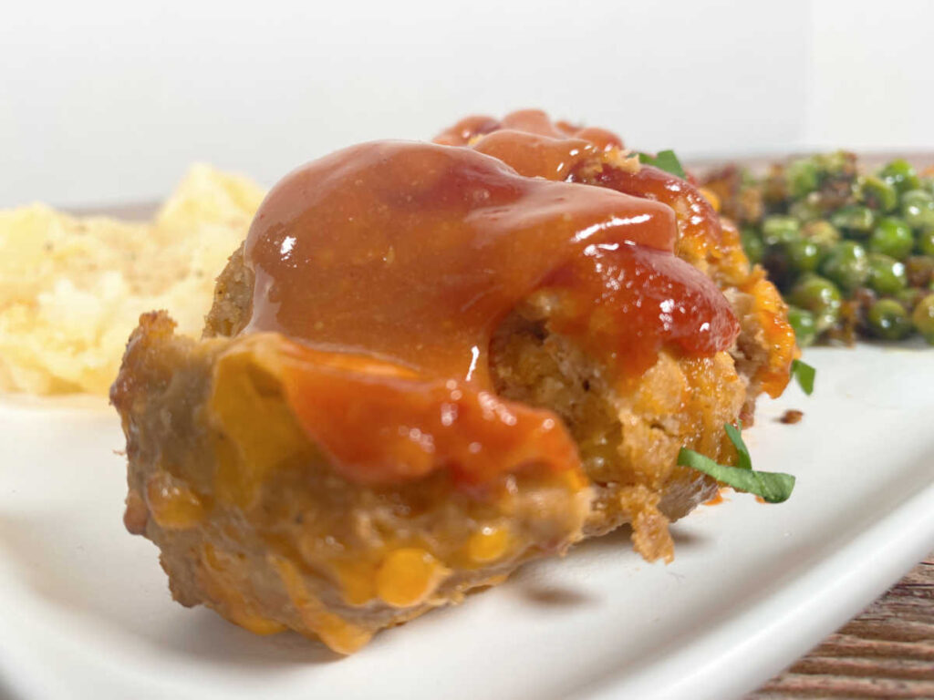 Side view of meatloaf sliced with the glaze on top. Mashed potatoes and peas are in the background of the plate.