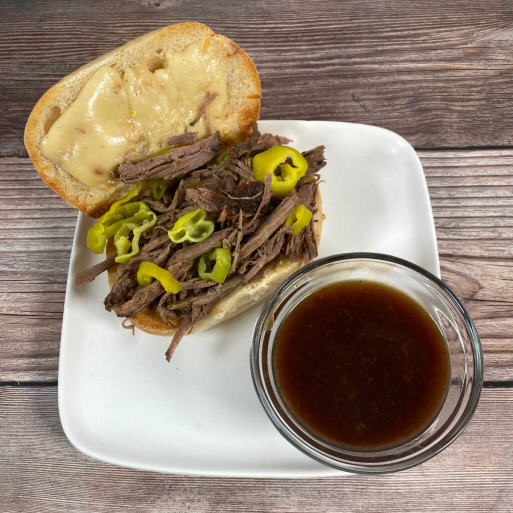 Process shot, plating - open sandwich with meat and pepperoncinis piled high, melted cheese on the bread and a dipping bowl for the gravy sit on a white plate. 