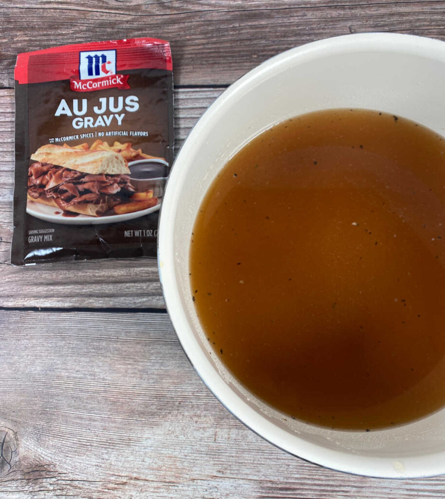 Process shot - packet of au jus gravy sits next to a saucepan with liquid from cooking the beef and water. 