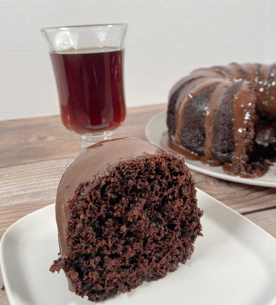 Close up image of slice of cake with whole cake and glass of coffee in the background. 