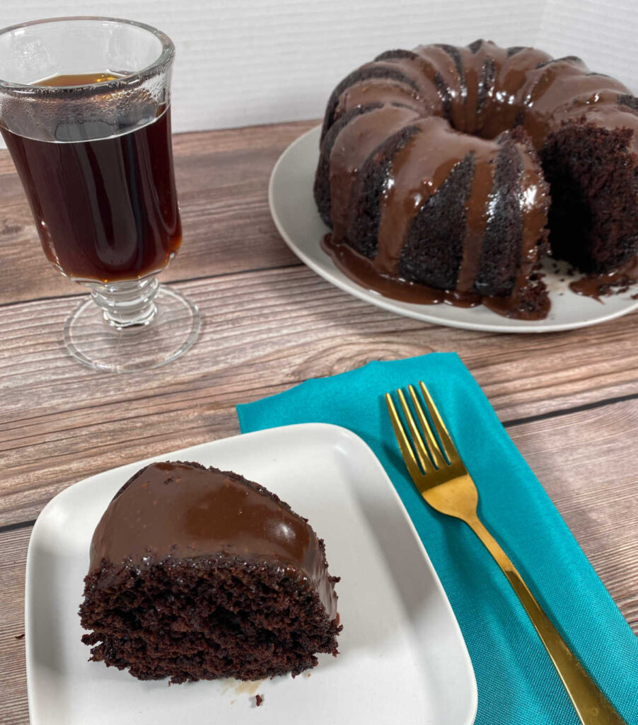 Close up image of frosted cake on plate with teal napkin and gold fork. Remainder of cake and glass of coffee in background. 