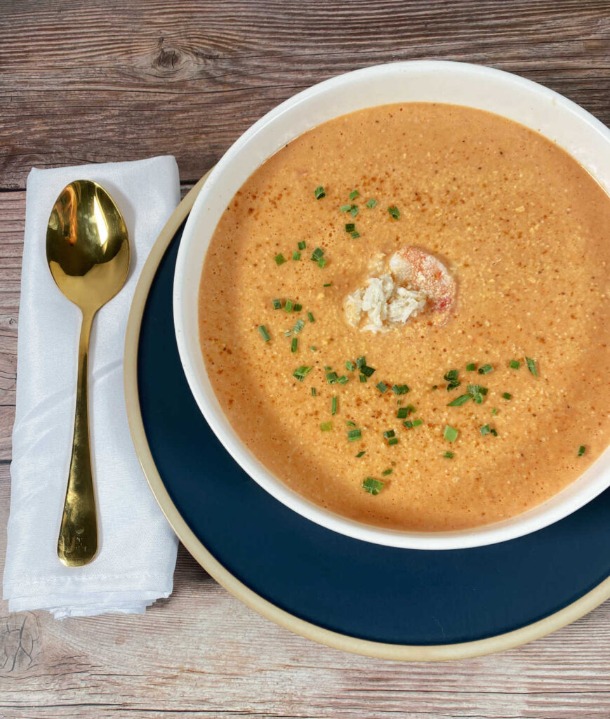 Soup in white shallow bowl, garnished with  shrimp and chives. Gold spoon on the left. 