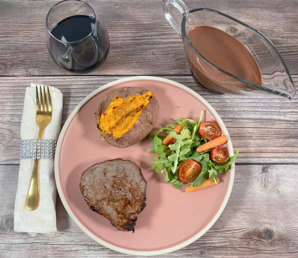 Steak and side dishes sit on a pink plate. Glass of white and chocolate sauce sits off to the side. 