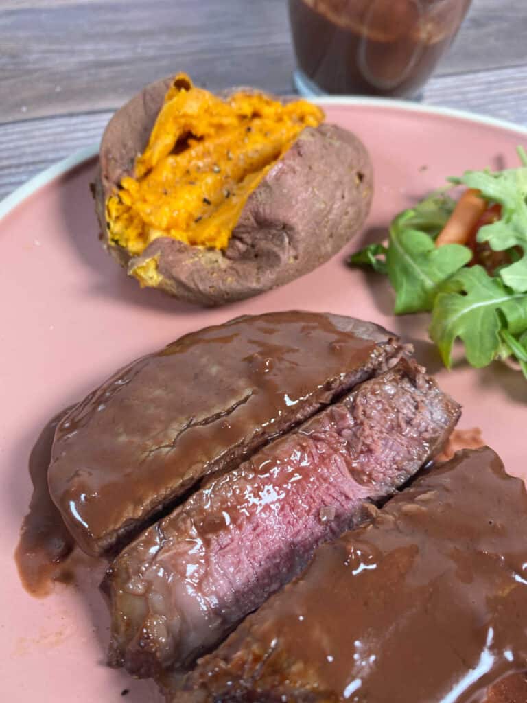 Slices of steak covered in sauce sit on a pale pink plate. 