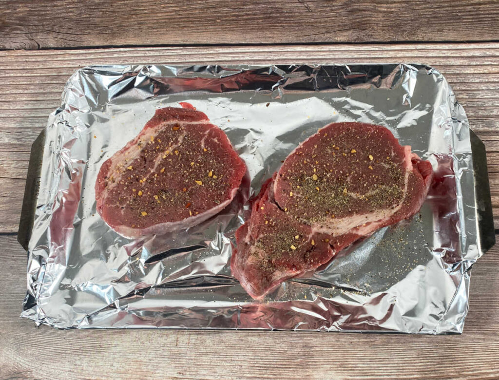 Process shot - seasoned steaks sit on a foil lined pan. 