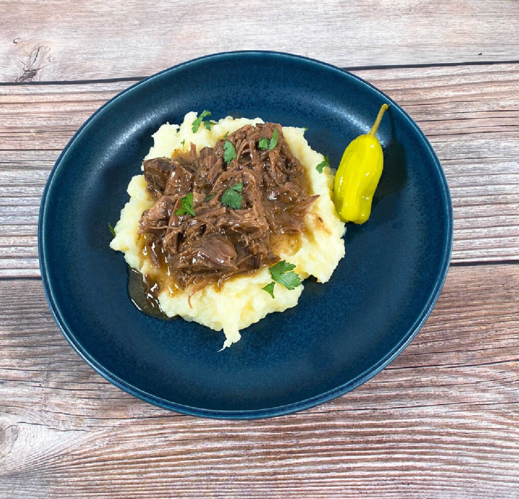 Pot Roast on top of mashed potatoes in a dark blue bowl. Garnished with parsley and a pepperoncini on the side.