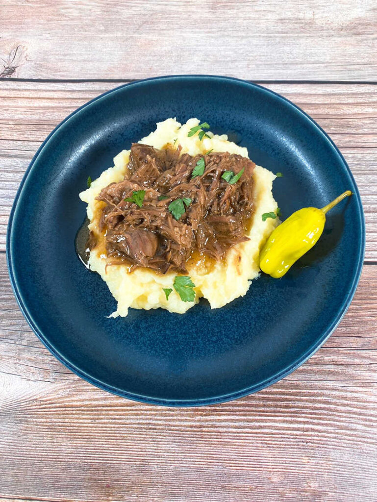 Pot Roast on top of mashed potatoes in a dark blue bowl. Garnished with parsley and a pepperoncini on the side.