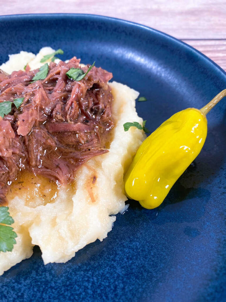 Close up image of pot roast on top of mashed potatoes in a navy blue bowl, with a pepperoncini on the side. 