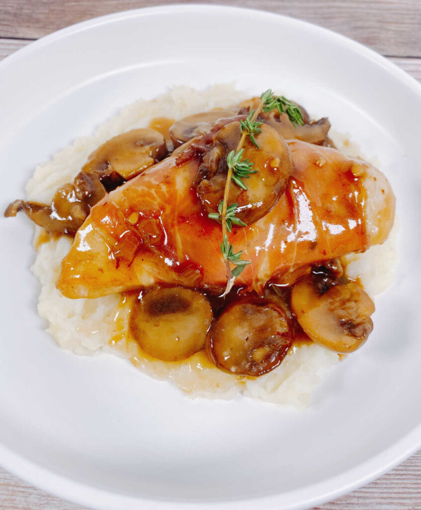 Recipe served over mashed potatoes in a round white dish. Mushrooms are under chicken and chicken is topped with a fresh sprig of thyme. 