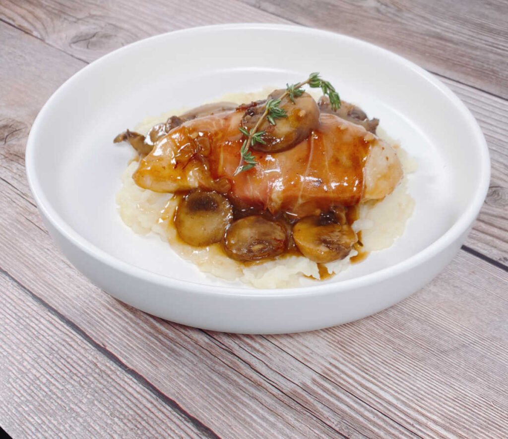 Prosciutto wrapped chicken sits on top the mushroom marsala sauce over mashed potatoes in a shallow white bowl. 