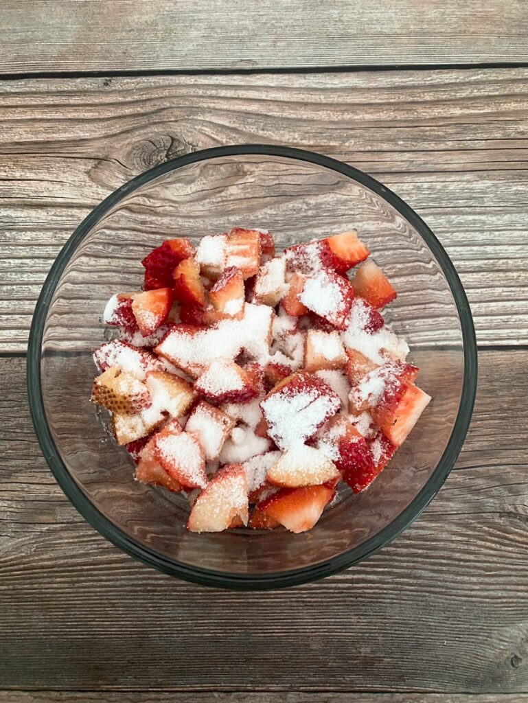 Process shot - diced strawberries in a glass bowl topped with sugar. 