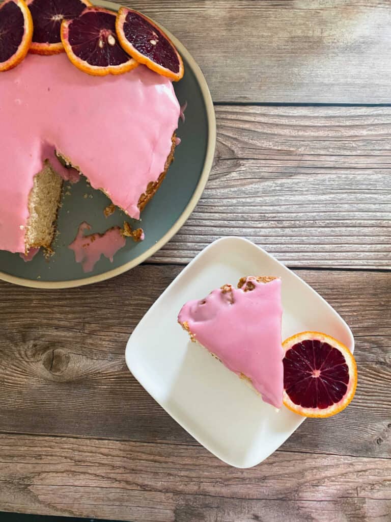 Slice of cake, garnished with blood orange slice, sits on a white square plate. 