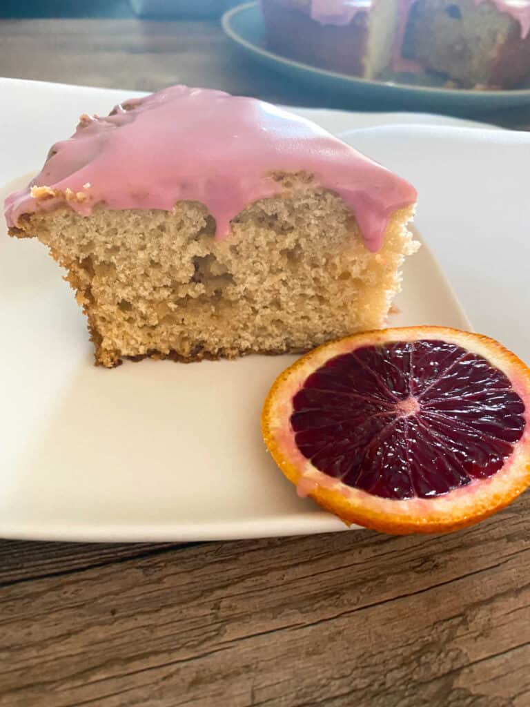 Slice of cake sitting on a white square plate, garnished with a blood orange slice. 