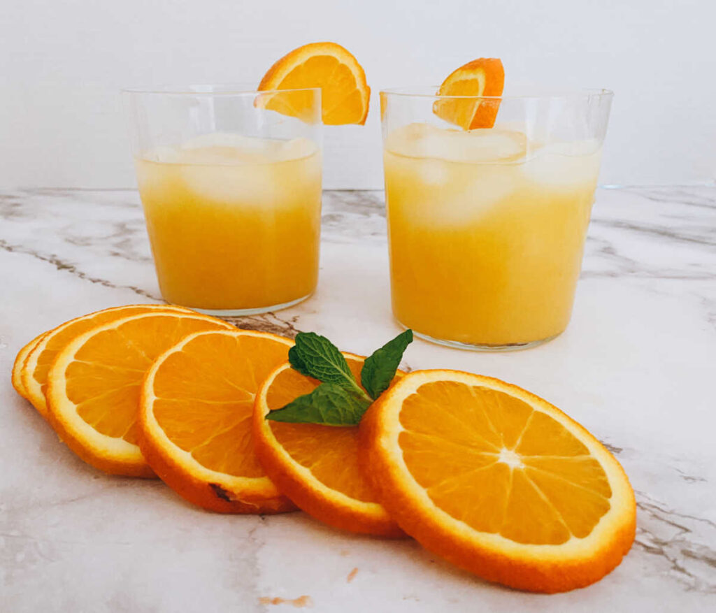 two glasses of the cocktail on a marble background with orange slices in the foreground.