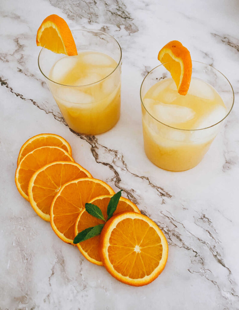 two glasses of the cocktail, garnished with orange wedges, on a marble background. 