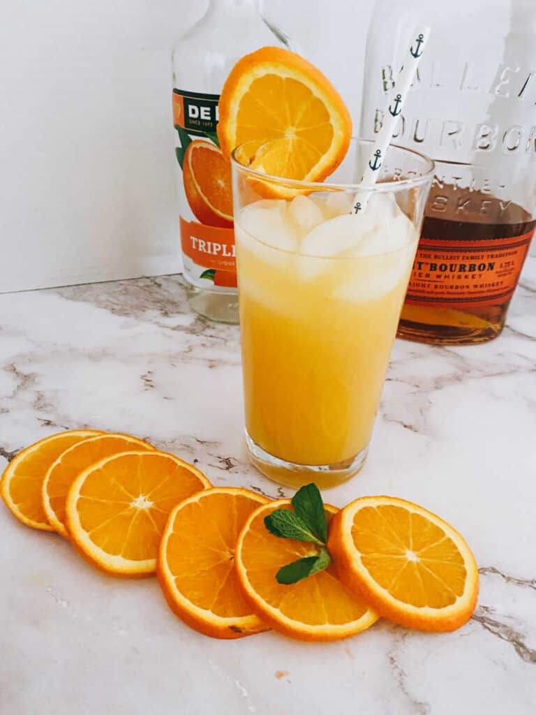 cocktail in glass, with liquor bottles in background and orange slices in foreground on a marble slab. 