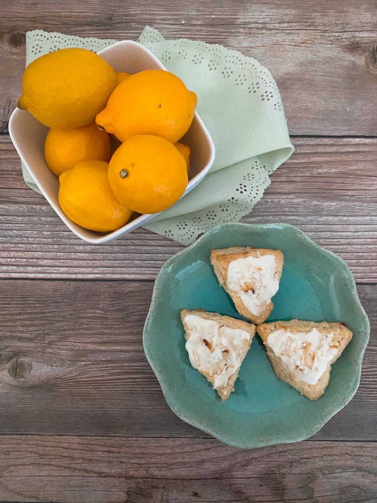 Three scones on a scalloped plate sit on a wooden background with a white bowl of lemons in the background. 