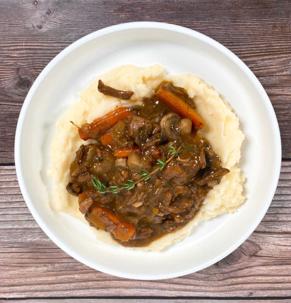 Stew, served over mashed potatoes, and garnished with a sprig of fresh thyme, sits in a shallow white bowl on a wooden background. 