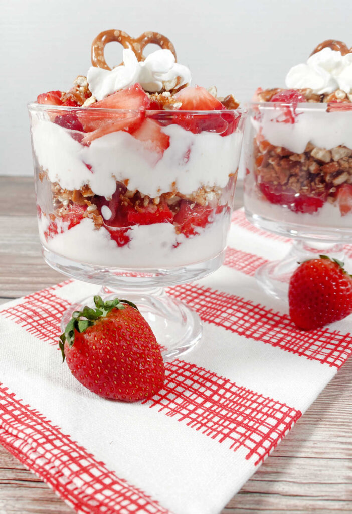 Two parfaits sitting on a red and white plaid towel, with strawberries in the front. 