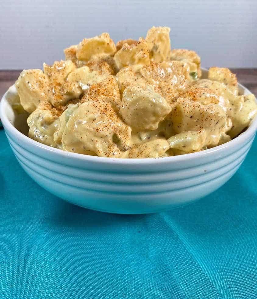 potato salad, garnished with paprika, sits in a white bowl on a blue napkin.