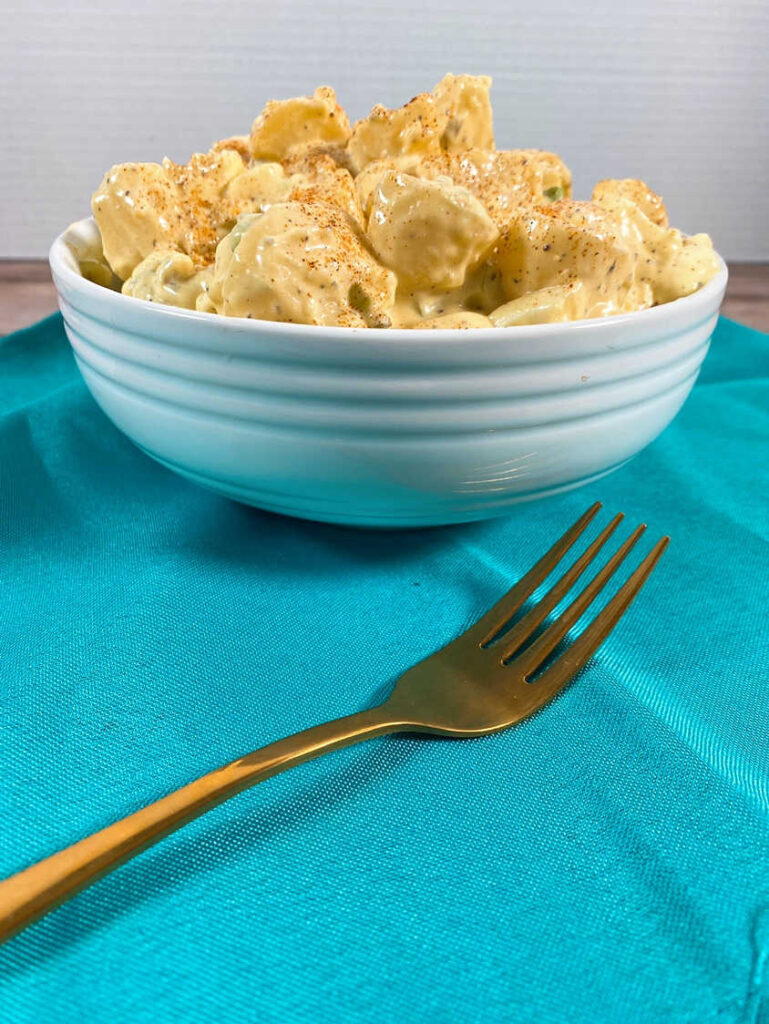 white bowl piled high with potato salad sits on a blue napkin. 