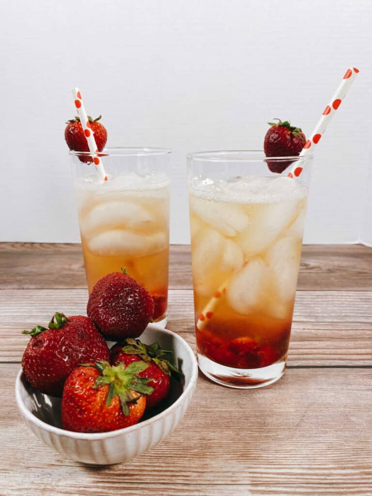 two cocktails sit on a wooden background with a white bowl of strawberries in front. 