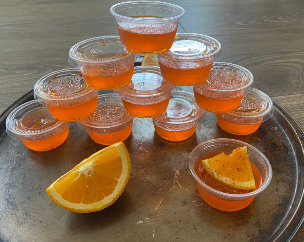 Pyramid of jello shots sits on a round pan, with an orange slice and an opened jello shot in front of them.