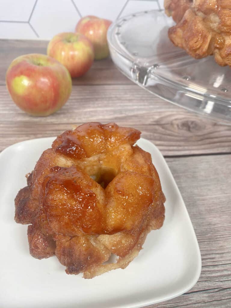 Monkeybread on a white plate, with other desserts on a glass cake stand and apples in the background. 