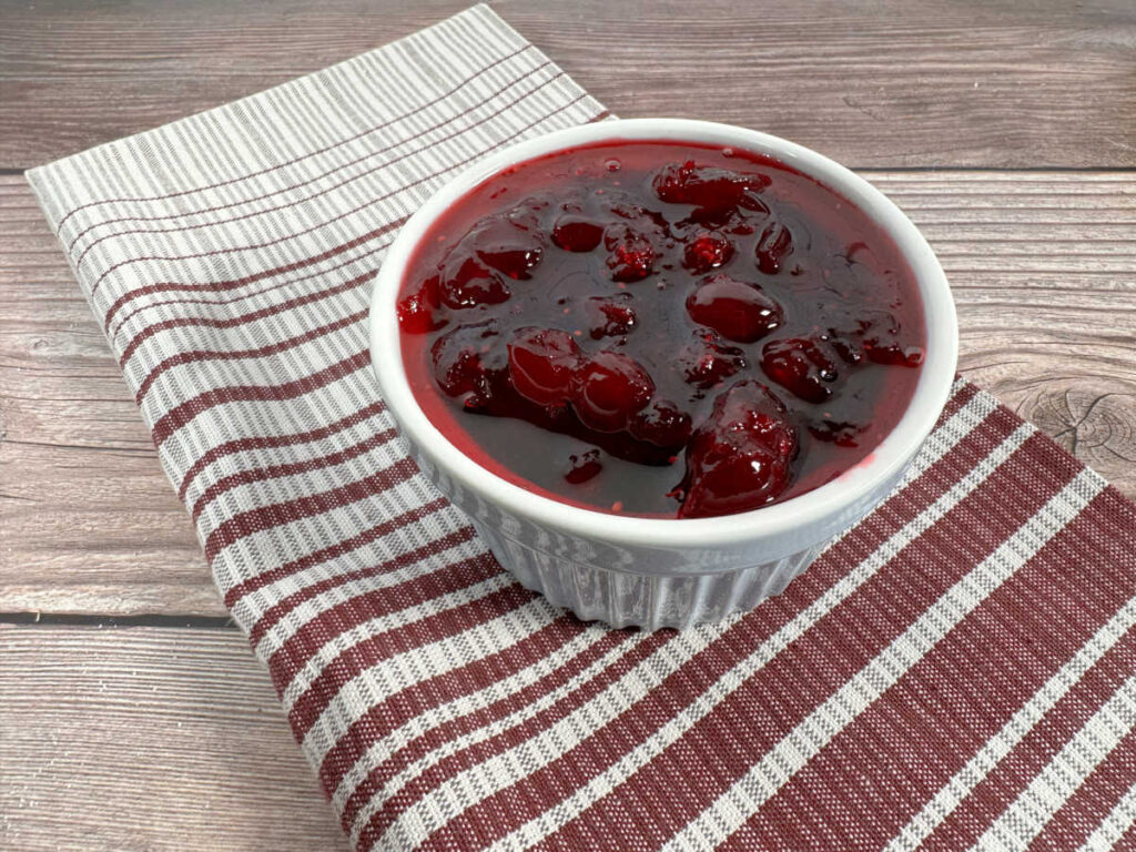 Cranberry sauce sits in a white ramekin on top a gray and burgandy striped cloth napkin. 