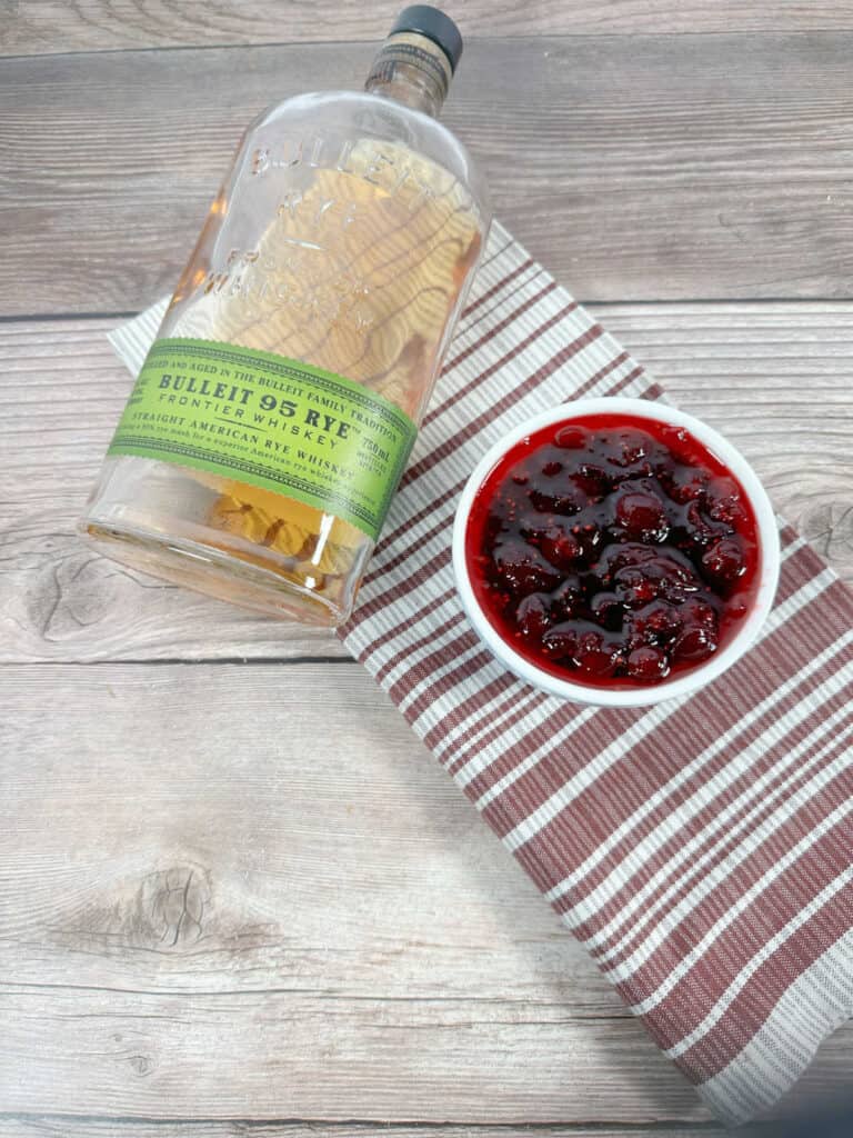 White ramekin filled with cranberry sauce sits on a gray and burgandy striped cloth napkin with a bottle of bourbon in the background. 
