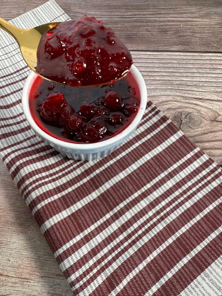 Cranberry sauce on a gold spoon hovers above a white ramekin of the sauce on a gray and burgandy striped napkin.