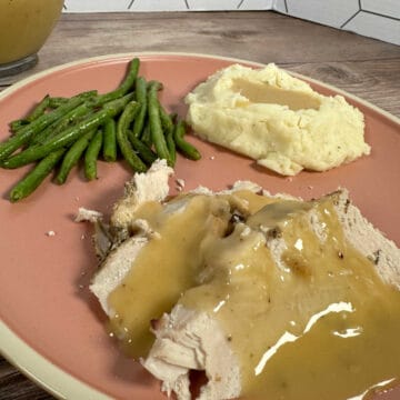 Turkey breast with gravy on a pink plate with sides of mashed potatoes and green beans.