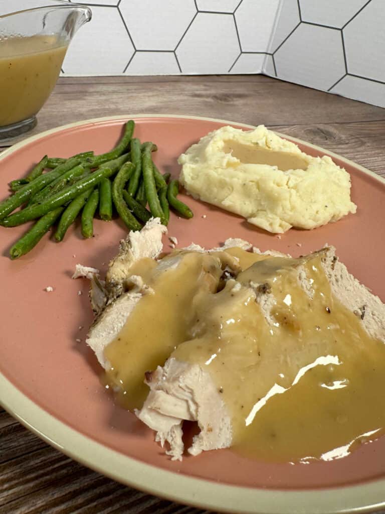 Turkey breast with gravy on a pink plate with sides of mashed potatoes and green beans.