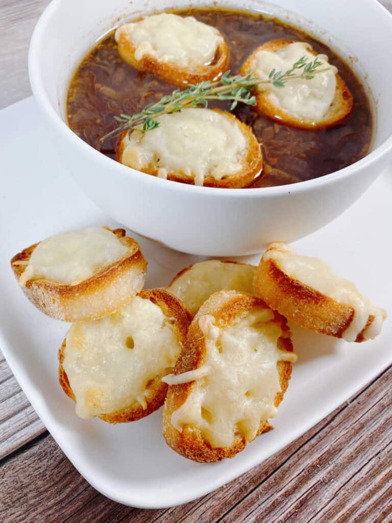 Soup in a white bowl on a white plate surrounded by pieces of cheesy toast. 