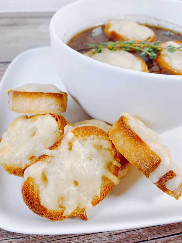 bowl of soup, topped with cheesy toast sits on a white plate with additional cheesy toast slices. 