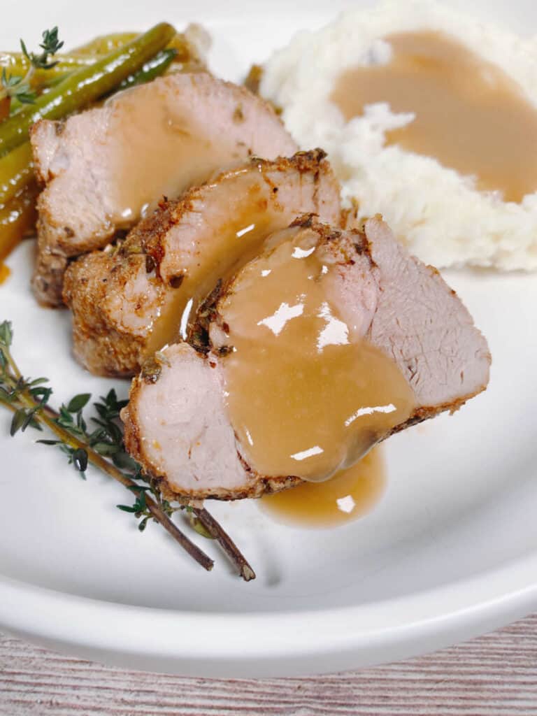 Close up of sliced pork, topped with gravy on a plate with mashed potatoes, green beans and fesh herbs. 