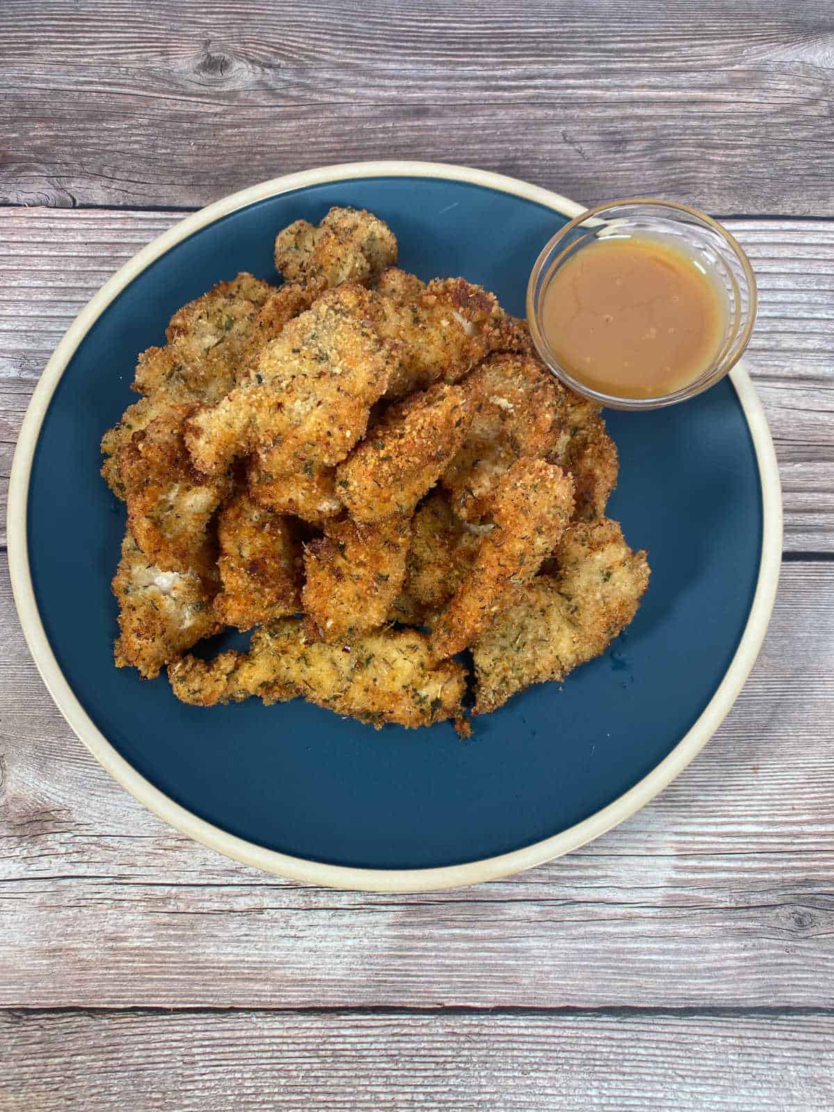 Overhead image of tenders piled high on a dark blue plate with a glass dish of honey mustard for dipping. 