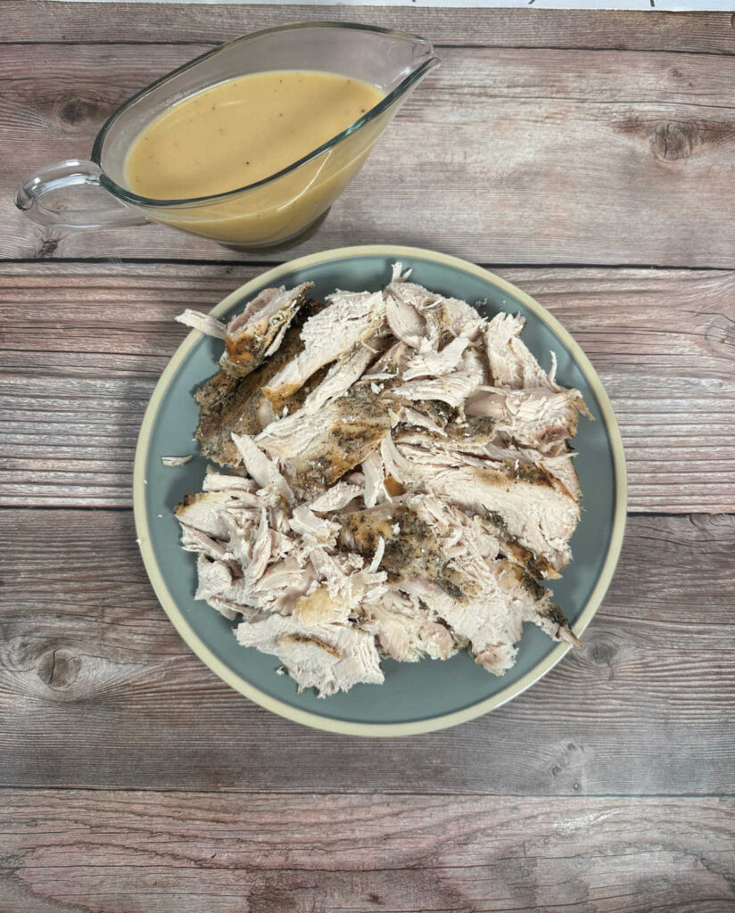 Overhead image of a round blue plate covered in sliced turkey breast on a wooden background. A full gravy boat sits behind it. 