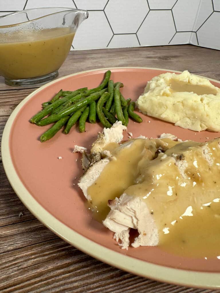 Round pink dinner plate with turkey breast and gravy, mashed potatoes and green beans on a wooden background. 