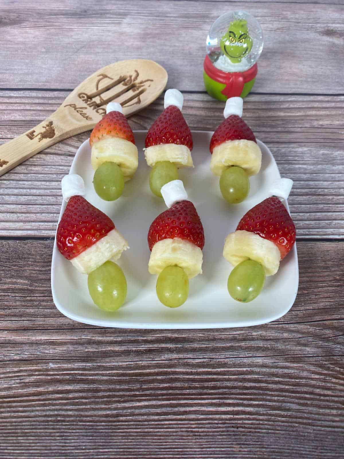 Skewers sit on a plate with a Grinch spoon and mini Grinch snow globe.
