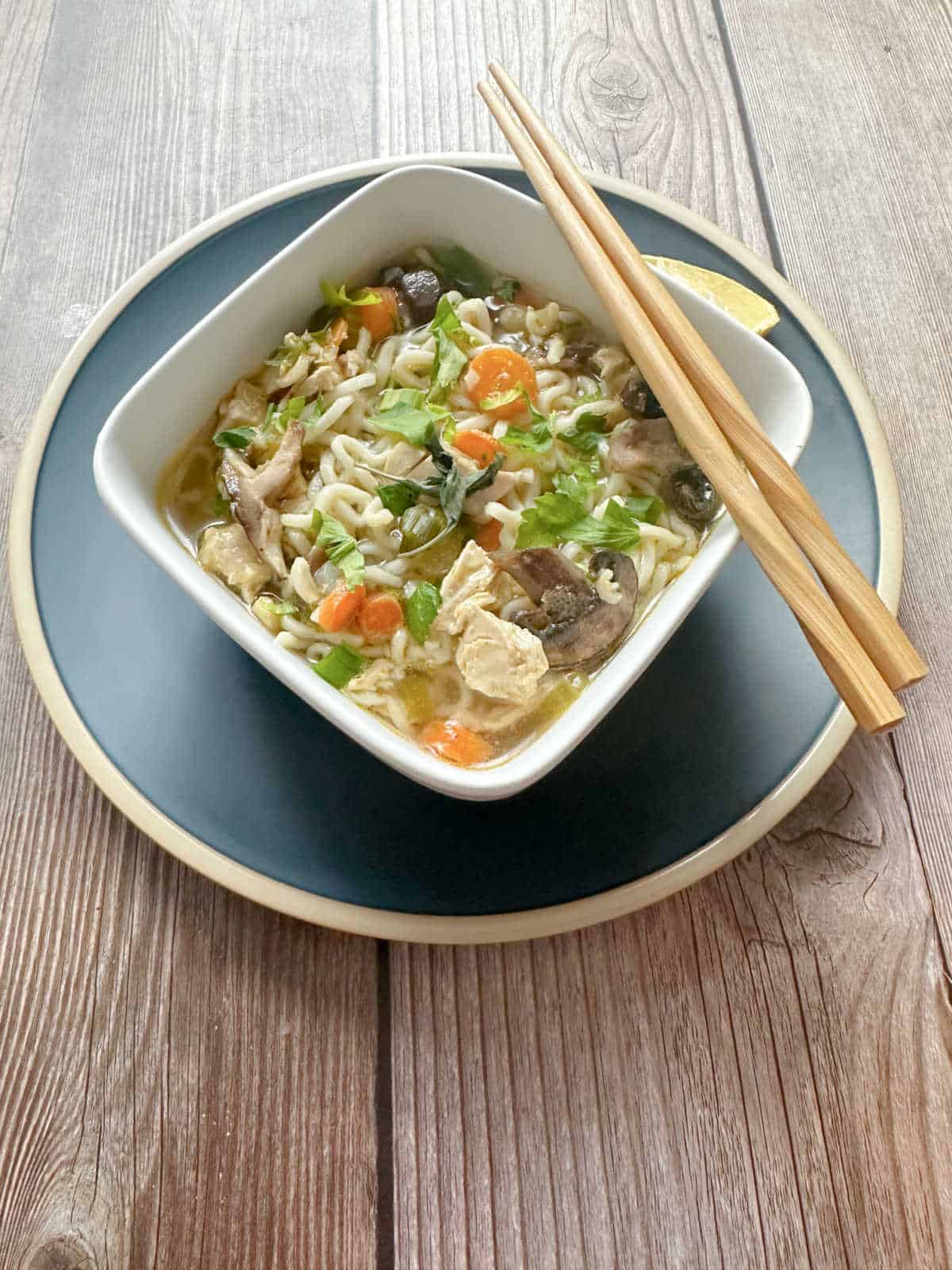 Soup in a square, white bowl on a blue plate with chopsticks resting on the side of the bowl.