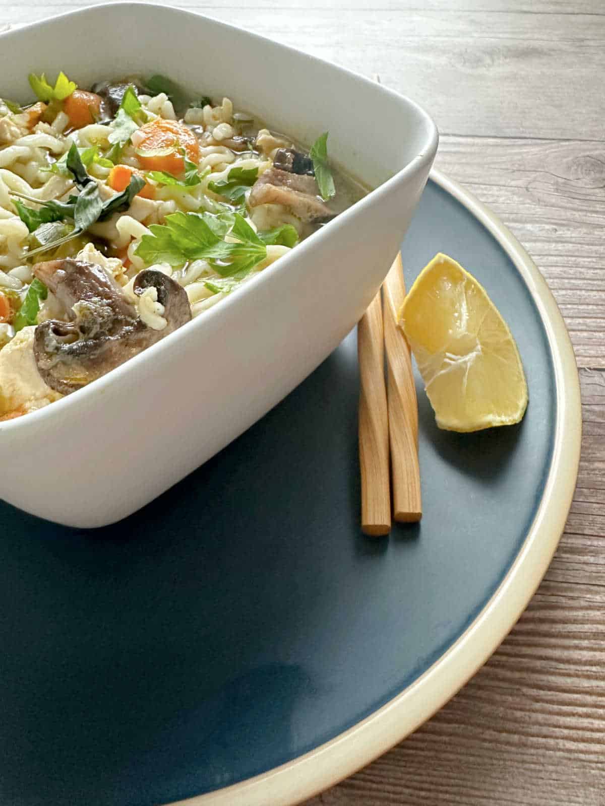 Close up image of soup in a square white bowl on a blue plate with chopsticks and a lemon wedge.