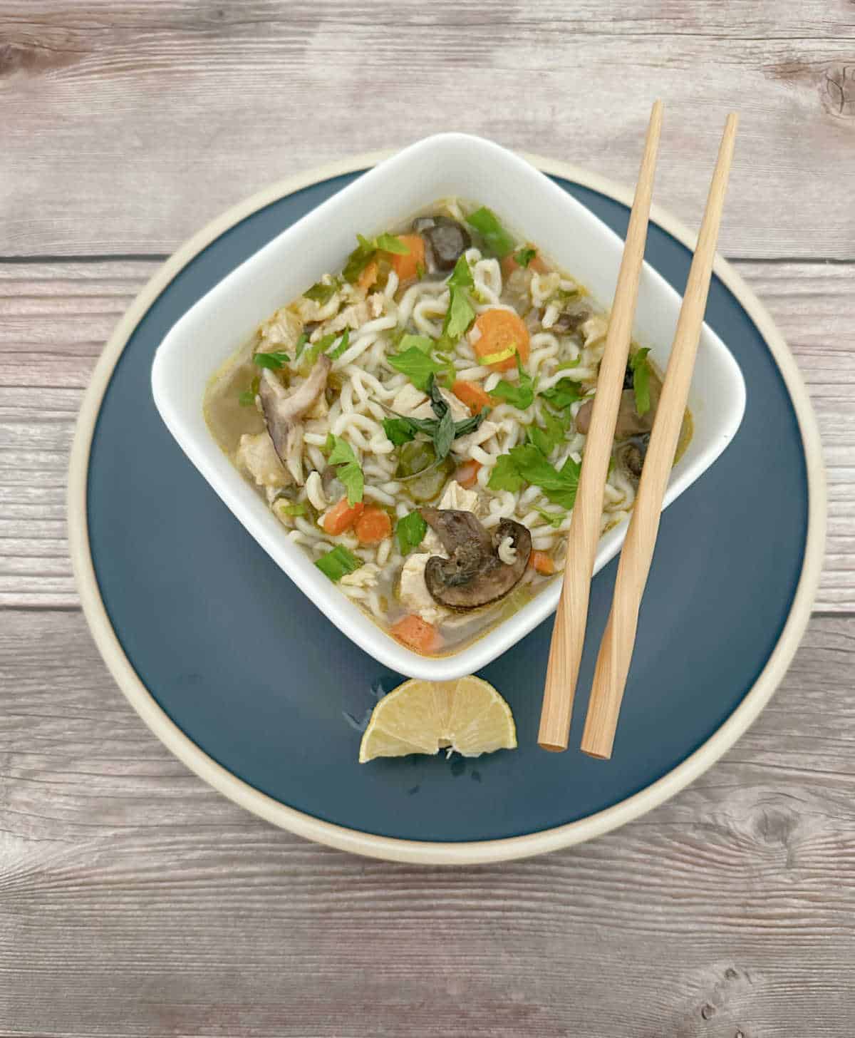 Soup in a square white bowl on a blue plate. Chopsticks sit on top of the bowl and a lemon wedge sits on the plate.