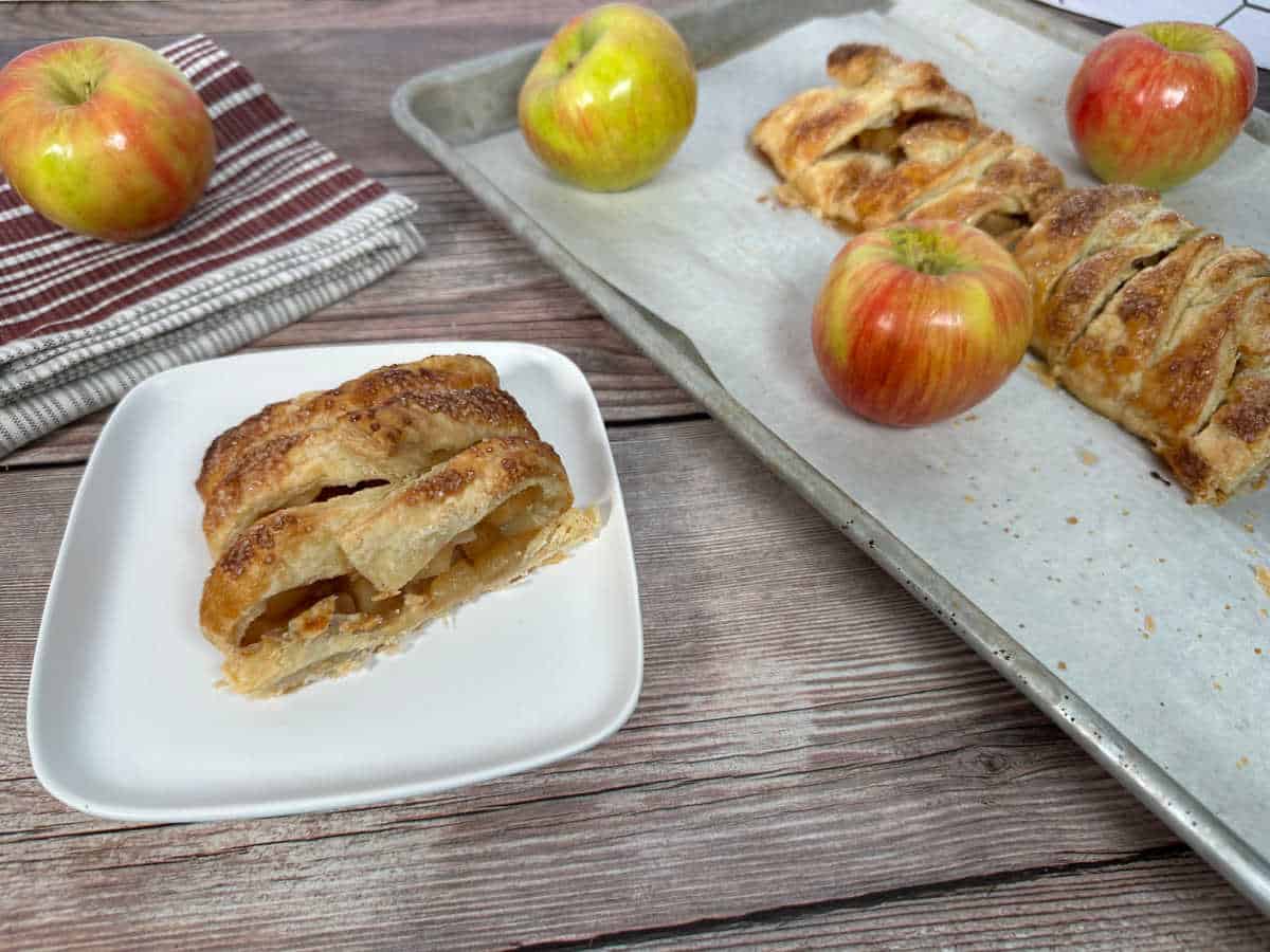 Slice of pastry on square white plate with baking sheet of remaining pastry off to the side. 