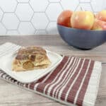 Slice of pastry sits on a red striped napkin on a wooden background. A blue bowl of apples is in the background.