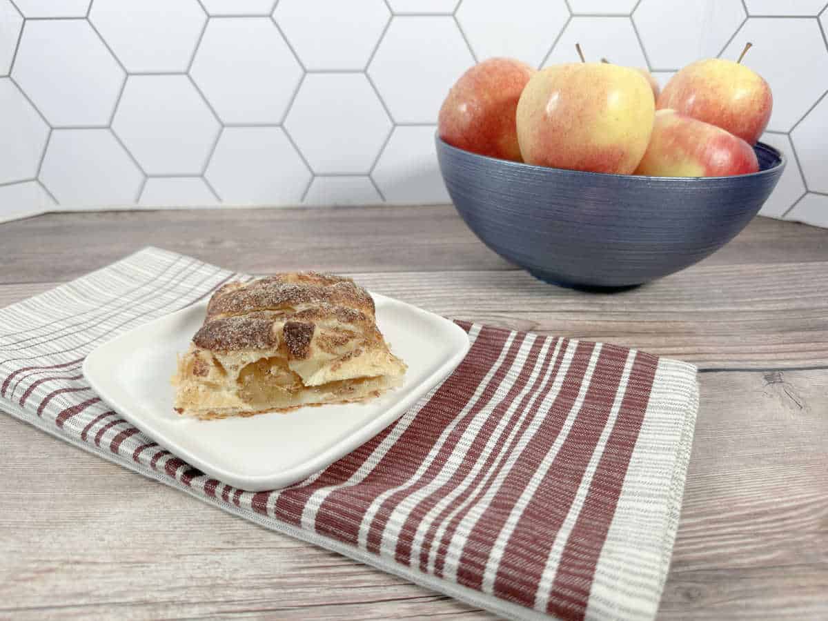 Slice of pastry sits on a red striped napkin on a wooden background. A blue bowl of apples is in the background.