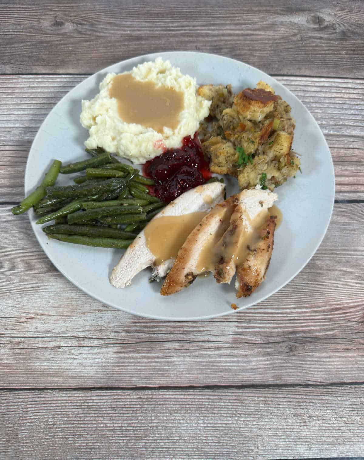 Slices of turkey breast, topped with gravy sit on a light blue plate surrounded by traditional Thanksgiving side dishes. 