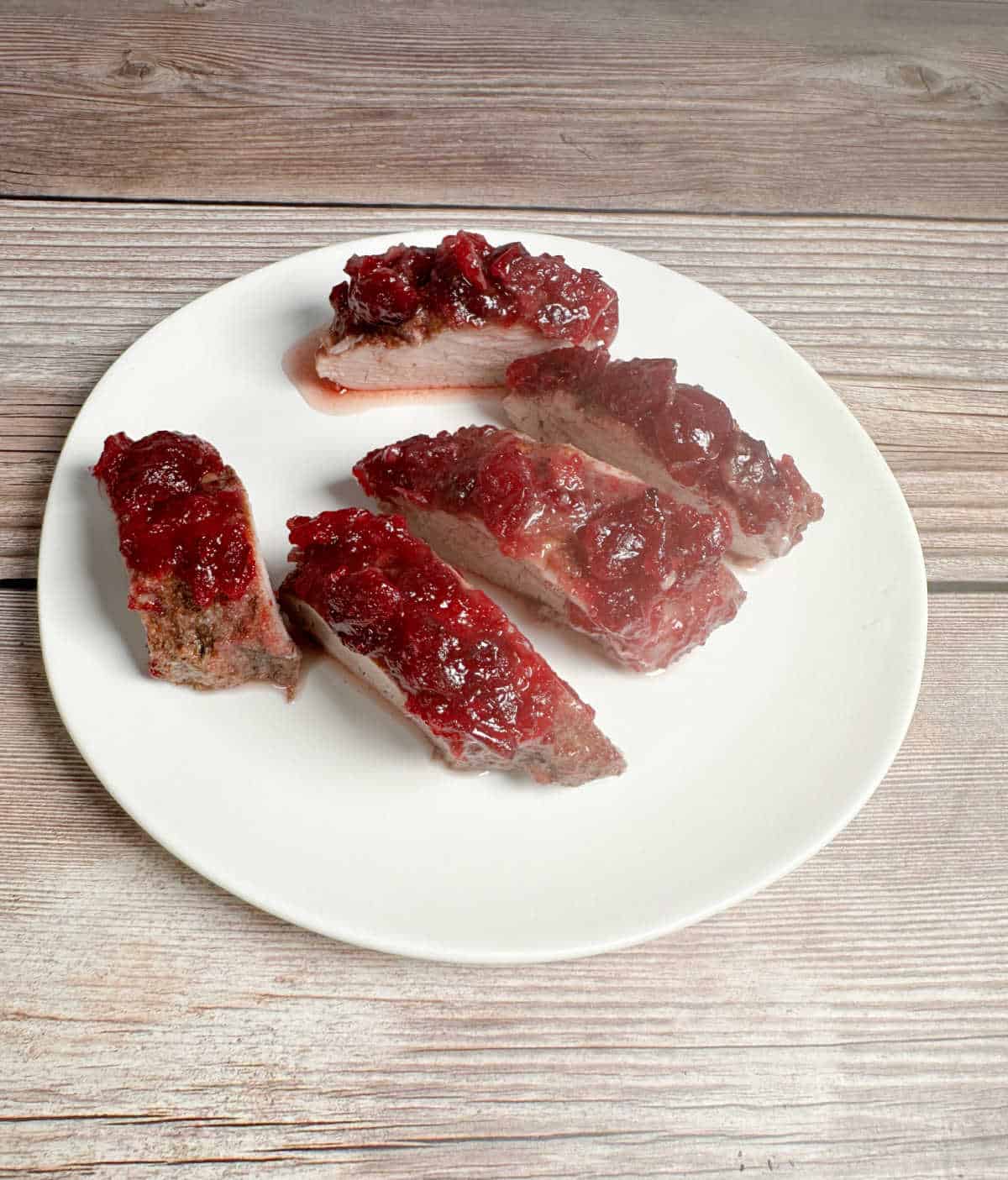 Slices of pork tenderloin topped with cranberry sauce sit on a round, white plate.